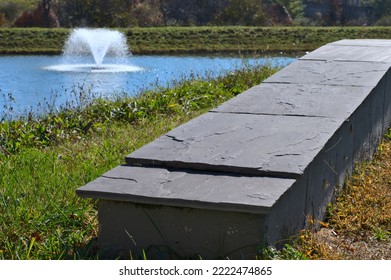 Shale Like Wall Barrier Between A Pond And A Sidewalk. The Small Wall Is A Dark Gray Color. The Pond Features A Water Fountain.