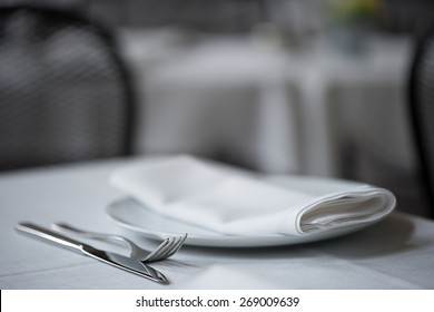 Shallow Depth Of Field Close Up Of Knife, Fork, Plate And Folded Napkin Laid Upon White Table Cloth At Elegant Restaurant.   