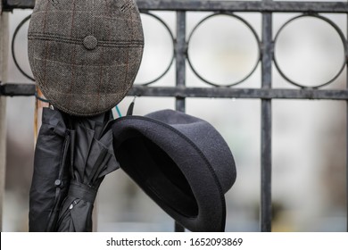 Shallow Depth Of Field (selective Focus) Image With Two Men's Hats And Two Black Umbrellas Hung On A Fence During A Rainy Day.