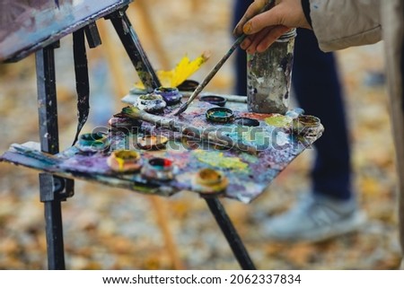 Shallow depth of field (selective focus) image with the hand of a female painter and her color palette while painting a landscape on a sunny autumn day.