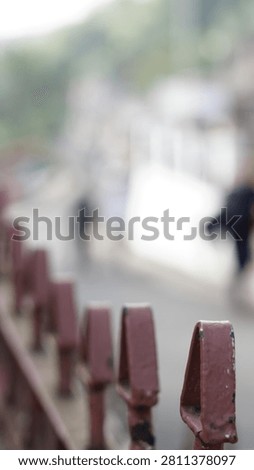 A shallow depth of field shot shows a rusty metal railing in the foreground with blurred figures walking in the background on a path.