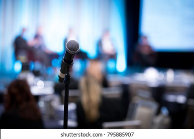 Shallow Depth Of Field View Of A Microphone On A Stand, With Blurry Panel Of Guest Speakers On Stage, At A Shareholder Conference