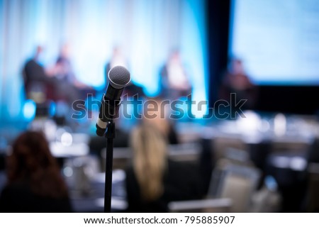 Shallow depth of field view of a microphone on a stand, with blurry panel of guest speakers on stage, at a shareholder conference