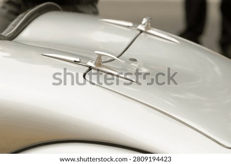 Shallow depth-of-field close view of a polished silver vintage car hood featuring twin streamlined chrome hood ornaments and smooth curved body lines. Soft