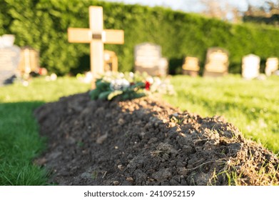 Shallow focus of a fresh burial showing some of the heaped soil on the grave site. A solitary wooden crucifix is at the head of the burial.