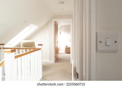 Shallow focus of a light switch seen at the exit to a bedroom. A large first floor hallway can be seen with skylight windows. In the distance is another first floor bedroom.