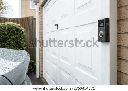 Shallow focus of a newly installed, wireless and rechargeable cloud-enabled doorbell attached to a down drain in a garage. A parked car can be seen on the wet, private driveway.
