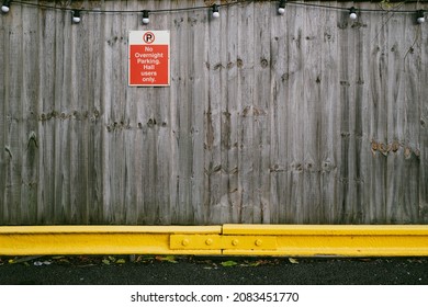 Shallow Focus On A Generic No Parking Sign Seen On A Wooden Fence Adjacent To A Village Community Centre. A Yellow Barrier Is Used To Protect The Privately Owned Garden Fence.