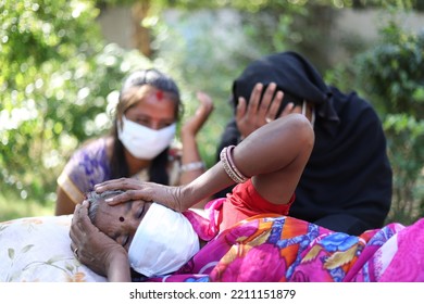 A Shallow Focus Shot Of An Old Indian Woman Wearing A Facemask, Lying Down In Pain With People Crying In The Background