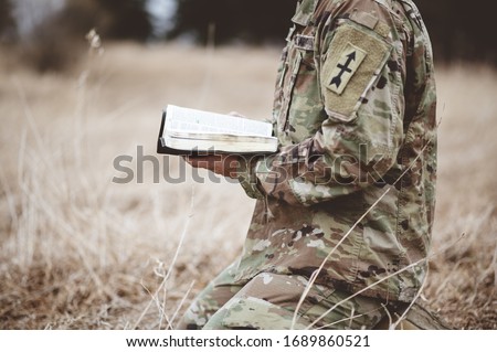 A shallow focus shot of a young soldier kneeling while holding an open bible in a field
