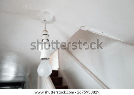 Shallow focus of a single electric light bulb hanging from the ceiling in a hallway of a Grade 2 listed farmhouse in Britain. The narrow stairs to the upstairs bedrooms can be seen.