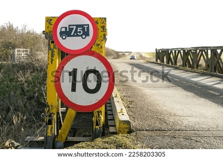 Shallow focus of a weight and speed restriction sign on a temporary bridge. A car can be seen in the distance, about to cross the fragile bridge.