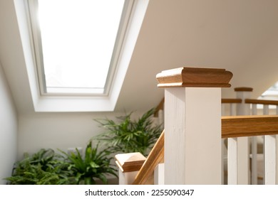 Shallow focus of a wooden bannister at the top of a recently converted loft space. Half way down the stairs are house plants on a ledge and nearby skylight window.
