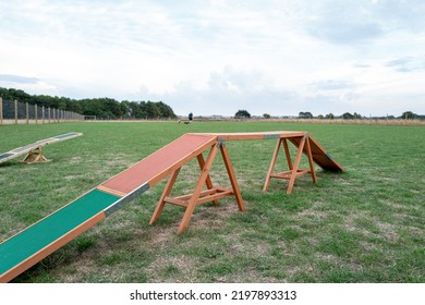 Shallow Focus Of A Wooden Dog Agility Sloping Bridge Seen In A Large Dog Agility Field. A Seesaw Is Just Visible And Dog With Owner The Background.