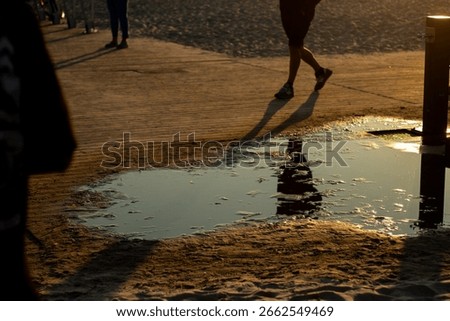 Shallow puddle on sandy beach path reflects golden sunset and walking figures, creating long dramatic shadows and textures.