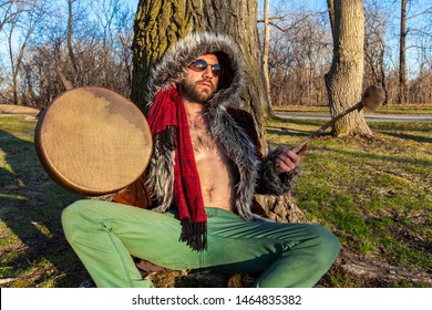 Shaman Plays Native Drum Under Tree. A Closeup Front View Of A Masculine Guy Wearing Green Trousers And Open Coat Revealing Bare Chest As He Plays Sacred Percussion Instrument And Relaxes In Nature