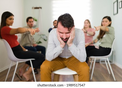 Shame And Guilt. Group Of People In A Circle Pointing To A Sad Latin Man. Depressed Young Man Feeling Judged For His Problems And Addictions