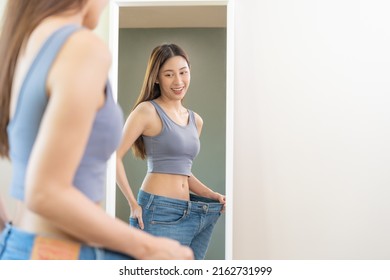 Shape slender, thin waist, attractive slim asian young woman, beautiful girl hand show shape her weight loss, wearing in big, large or over size jeans and looking into mirror. People body fit healthy. - Shutterstock ID 2162731999