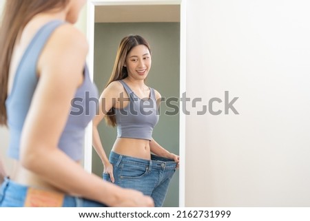 Shape slender, thin waist, attractive slim asian young woman, beautiful girl hand show shape her weight loss, wearing in big, large or over size jeans and looking into mirror. People body fit healthy.