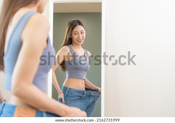 Shape slender, thin waist, attractive slim asian\
young woman, beautiful girl hand show shape her weight loss,\
wearing in big, large or over size jeans and looking into mirror.\
People body fit healthy.