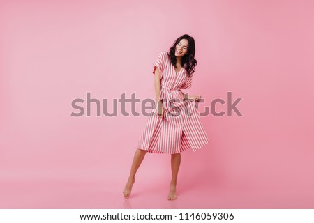 Shapely barefooted lady with tanned skin dancing on pink background. Happy caucasian girl in striped dress fooling around and laughing.