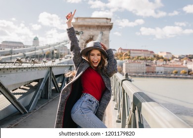 Shapely blissful girl in red sweater dancing on bridge on blur city background in autumn morning. Outdoor photo of happy female tourist having fun, exploring attractions. - Powered by Shutterstock