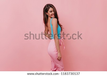Shapely lovable girl with beautiful smile posing in studio. Carefree brunette female model in romantic attire standing near pink wall.