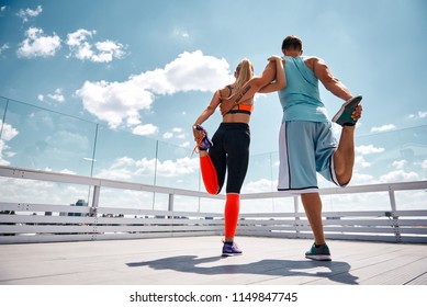 Shapely man and woman are exercising jointly on top of high building. They are standing on one leg and bending other. Partners are supporting each other by embracing while focus is on back