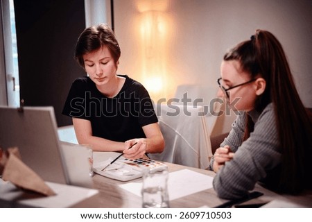 In a shared moment of creativity, one artist applies paint while her counterpart watches closely, both deep in the artistic zone amidst the cozy studio lights.