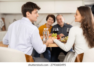 sharing close moment at dining table, holding hands and locking eyes, while happy parents watching in background at family home. Blessing of parents