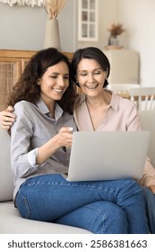 Sharing digital experience. Smiling older age mother millennial daughter cuddle on sofa relax with notebook device watch funny video look through family photos make purchases at online ecommerce store