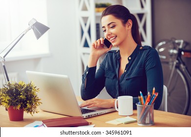 Sharing good business news. Attractive young woman talking on the mobile phone and smiling while sitting at her working place in office and looking at laptop