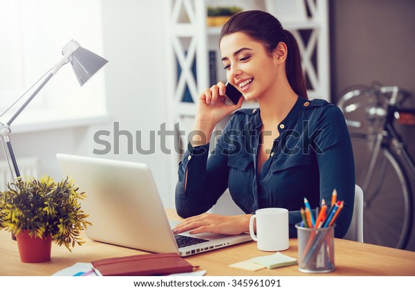 Sharing good business news. Attractive\
young woman talking on the mobile phone and smiling while sitting\
at her working place in office and looking at\
laptop
