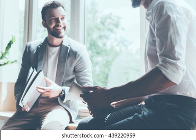 Sharing good news. Cropped image of two young businessmen in smart casual wear talking and smiling while sitting on the window sill in office  - Shutterstock ID 439220959