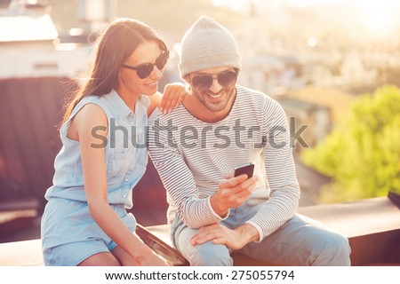 Sharing great pictures with friends. Beautiful young couple looking at the mobile phone together while sitting on the roof of the building