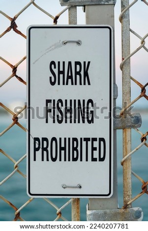 Shark Fishing Prohibited sign board at a pier with ocean view in Destin Florida. Close up of a black and white warning signage on a rusty and old chain link fence or gate.
