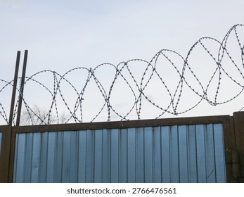 Sharp barbed wire stretched along top of blue corrugated metal security fence against overcast sky.