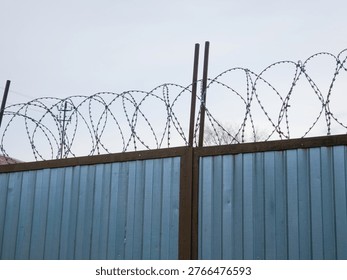 Sharp barbed wire stretched along top of blue corrugated metal security fence against overcast sky.