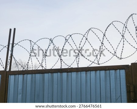 Sharp barbed wire stretched along top of blue corrugated metal security fence against overcast sky.