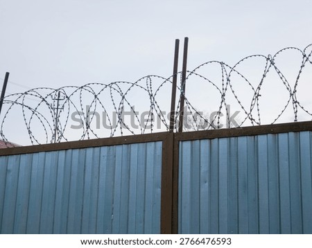 Sharp barbed wire stretched along top of blue corrugated metal security fence against overcast sky.