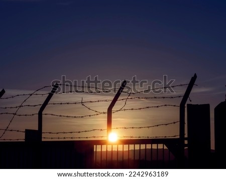 Sharp barber wire on a metal fence. Dark sunset cloudy sky in the background. Nobody. Security and asset protection. Defense perimeter. Sun flare. Dark and moody feel.