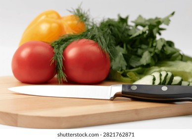 A sharp chef knife lies on a wooden cutting board surrounded by ripe tomatoes, sliced cucumber, yellow bell pepper and fresh herbs, suggesting home cooking, meal prep and healthy vegetable ingredients.