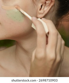 Sharp close-up of a hand using a wand applicator to precisely apply a stroke of liquid natural-tone foundation or concealer over a cheek area prepped with green color corrector