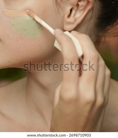 Sharp close-up of a hand using a wand applicator to precisely apply a stroke of liquid natural-tone foundation or concealer over a cheek area prepped with green color corrector