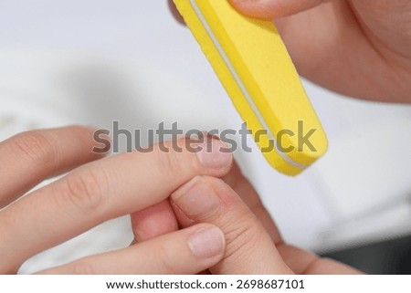 A sharp close-up shot capturing the action of a bright yellow foam nail buffer block being used by a manicurist to file and refine the rough edge and surface of a client's fingernail