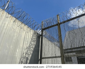 Sharp coiled razor wire lines the top of a formidable security fence and concrete wall, casting intricate shadows under a bright blue sky