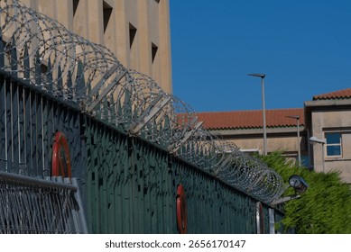 Sharp, coiled razor wire tops a tall fence, casting a stark, secure tone against a clear blue sky and a building with a red tiled roof.