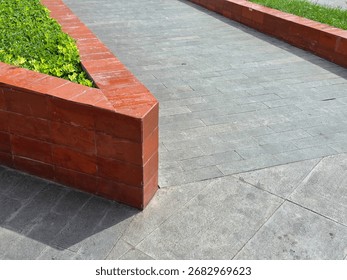 ​A sharp corner of a glossy, red-brick retaining wall for a planted area meets a textured gray tiled walkway under bright sunlight.