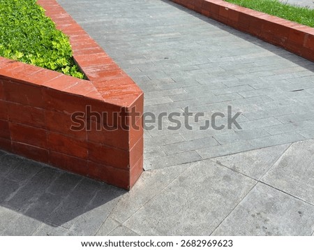 ​A sharp corner of a glossy, red-brick retaining wall for a planted area meets a textured gray tiled walkway under bright sunlight.
