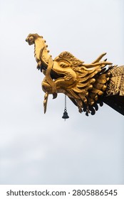 A sharp, detailed study of a gilded Makara gargoyle crowning the roof eave of a Tibetan Buddhist temple in Yunnan, China. The mythological sea creature is captured in stark profile against a pale, uniform sky, drawing immediate attention to the masterfully carved textures of its feathered gills, curling horn, and bared fangs. A delicate, weathered bronze wind bell hangs from its lower jaw, ready to catch the mountain breeze. The image exemplifies the transcendent metalwork and spiritual iconography that guards the sacred sanctuaries of the East.